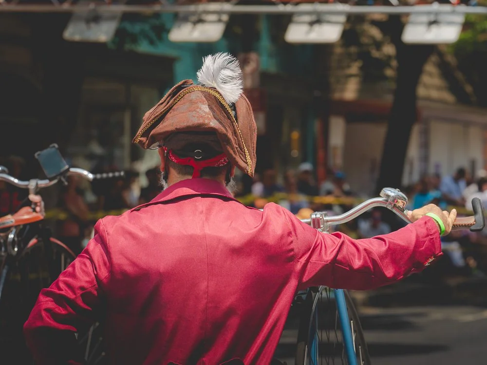 A cyclist in a red coat and feathered hat holding a high-wheel bicycle