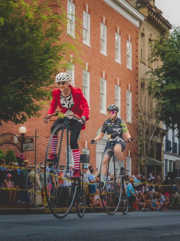 Two women riding high-wheel bicycles past Frederick's brick architecture