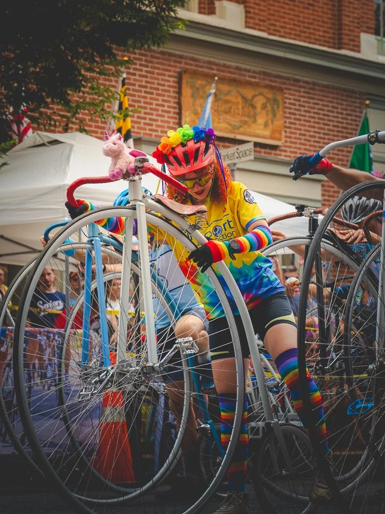 A rainbow-clad cyclist steadying a decorated high-wheel bicycle