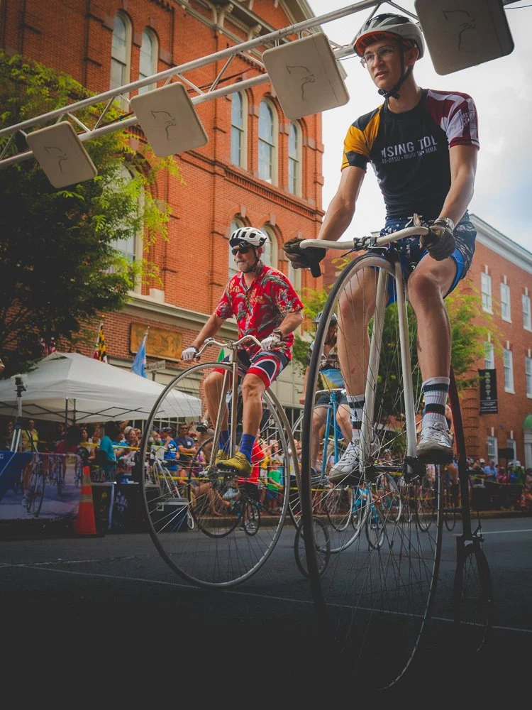 Two cyclists riding high-wheel bicycles past a brick building in Frederick