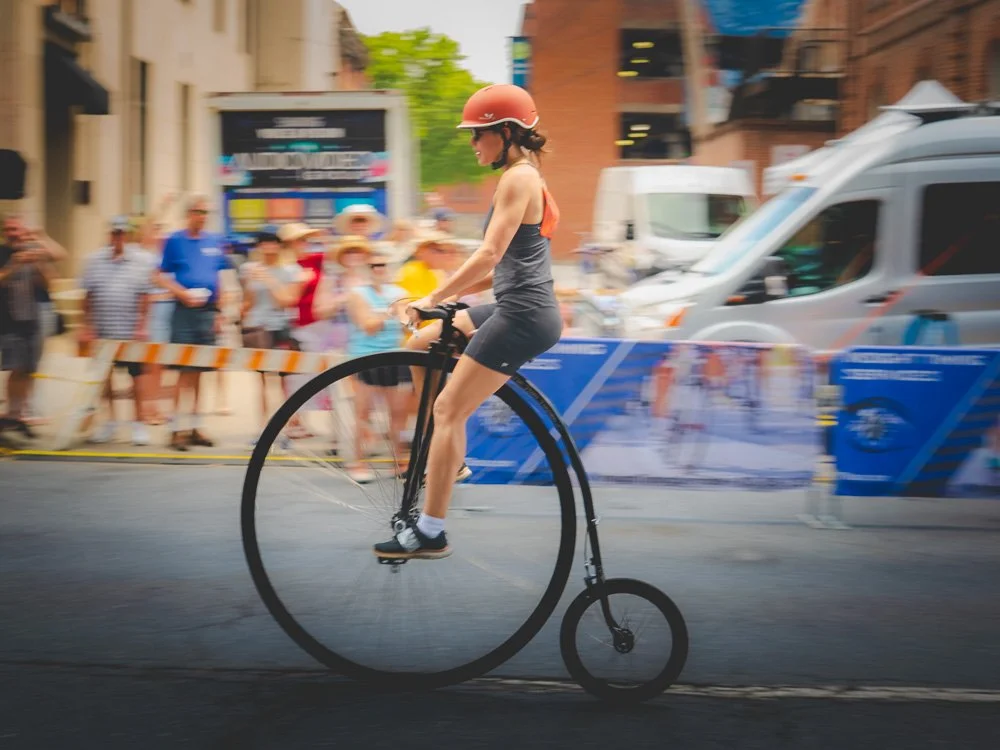 A high-wheel cyclist speeding past spectators with the city blurred behind her