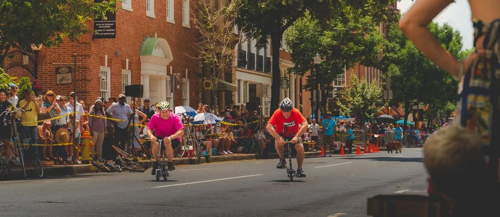 Two riders racing tiny bicycles as spectators line the street