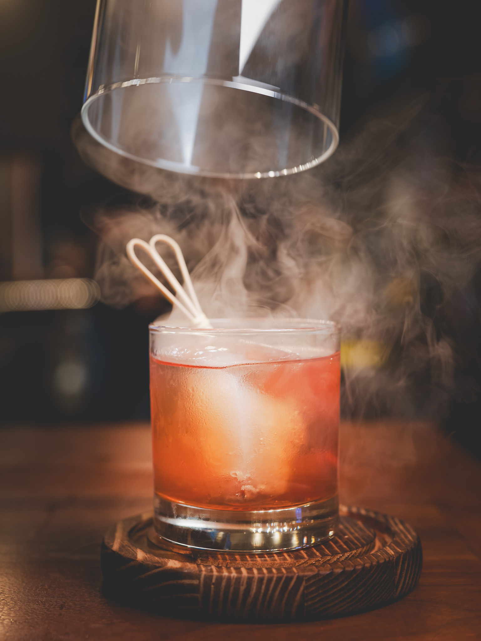 Smoke curling around a whiskey cocktail beneath a glass cloche